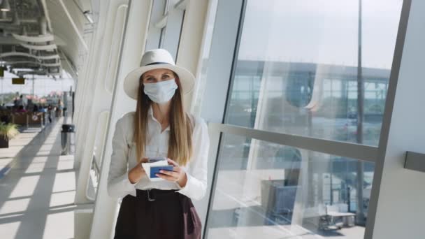 Hermosa mujer de negocios en el aeropuerto, utilizando el teléfono para enviar mensajes de texto, de pie junto a la ventana en el edificio de oficinas corporativas, mirando a la cámara. La chica tiene entradas y... — Vídeos de Stock