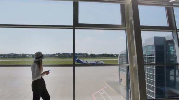 Молодая женщина путешественник, досуг или бизнес стоя у стеклянного окна крупного международного аэропорта хаба с телефоном в руках, прокручивая приложение для смартфонов. Фон самолета — стоковое видео