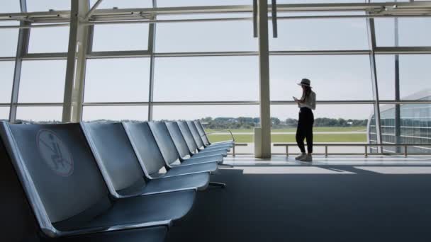 Mulher viajante, lazer ou negócios em pé na janela de vidro de um grande hub aeroporto internacional com um telefone em suas mãos, rolando o aplicativo para smartphones. Nas mãos dos bilhetes — Vídeo de Stock