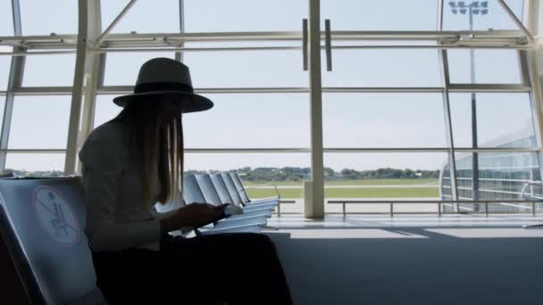 Menina de negócios elegante bonito irreconhecível sentado no aeroporto, segurando um telefone e bilhetes, aproximando-se lentamente da janela do aeroporto. Passarela do avião em segundo plano — Vídeo de Stock