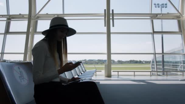 Elegancka, piękna, nierozpoznawalna bizneswoman siedząca na lotnisku, trzymająca telefon i bilety, powoli podchodząca do okna lotniska. Pas startowy samolotu w tle — Wideo stockowe