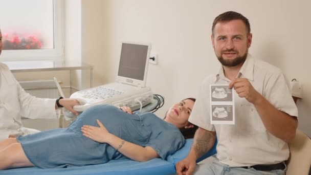 O pai tem uma foto do feto nas mãos. Retrato de um pai feliz. No fundo encontra-se uma mulher grávida — Vídeo de Stock