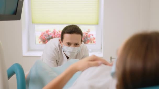 Ginecologista experiente em jaleco, máscara facial e luvas segurando o espéculo vaginal médico para examinar o paciente. Jovem deitada na cadeira ginecológica durante o check-up. — Vídeo de Stock