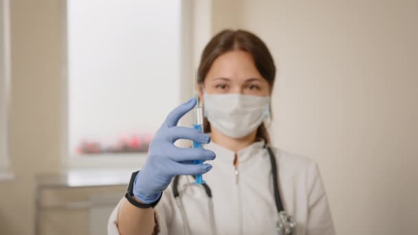 Female doctor with medical mask and gloves prepares prefilled syringe for injection by removing cap, tapping the syringe, ejecting a few drops out of the hypodermic needle. Vaccination, immunization. — Stock Video