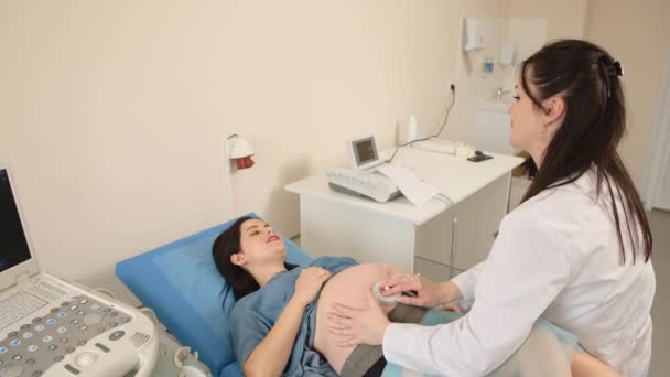 Vista superior de la mujer bastante embarazada acostada en el sofá médico mientras que el ginecólogo competente examinar los latidos del corazón del bebé. Equipamiento moderno para chequeo. Atención médica durante el embarazo. — Vídeo de stock