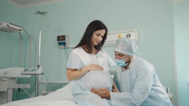 Gondoskodó fehér férfi orvosi egyenruhában, aki a terhes feleségét támogatja a kórházi kórteremben. Boldog nő nézi a férjét és fogja a kezét. Szülés partner koncepcióval. — Stock videók