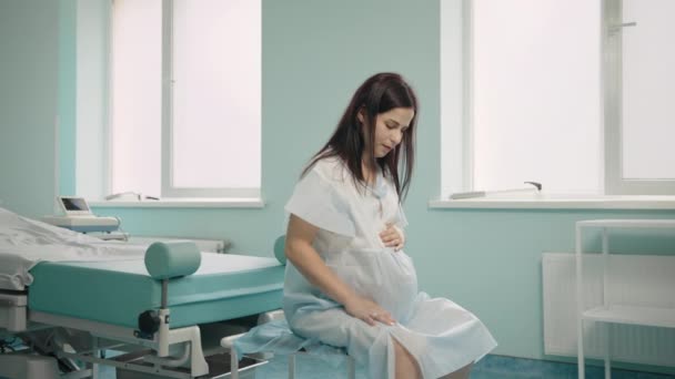 Mujer embarazada caucásica con contracción dolorosa sentada en la sala del hospital, tocando el vientre y mirando hacia abajo. Proceso de parto en el hospital. — Vídeos de Stock