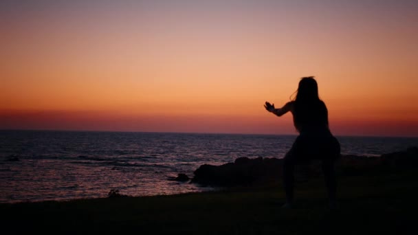 Fitness žena v siluetě dělá dřepy cvičení na pláži u oceánu. Mladá žena ve sportovním oblečení tráví večer aktivitami venku. Úžasný barevný západ slunce na pozadí. Fitness žena — Stock video