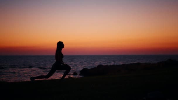 Активна жінка розтягує тіло під час заходу сонця над океаном — стокове відео
