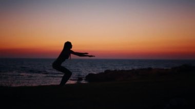 Silüetli kadın, okyanus kıyısındaki sahilde çömelme egzersizi yapıyor. Spor giyim sektöründe genç bir bayan akşam vaktini dışarıda geçiriyor. Arka planda inanılmaz renkli günbatımı. Fitness kadını