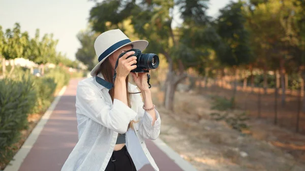 Gelukkige jonge vrouw met behulp van moderne digitale camera voor het nemen van foto 's tijdens het lopen. Vrouwelijke toerist in zomerhoed en kleding die herinneringen van de reis bespaart. Jonge vrouw fotograaf zwerver — Stockfoto