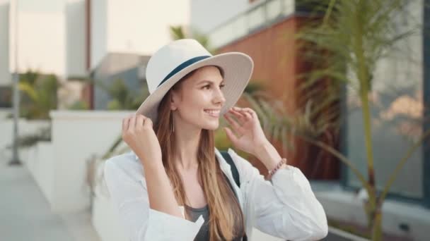 Mujer joven positiva con ropa elegante y sombrero blanco sonriendo sinceramente en la cámara mientras está de pie en la calle. Clima soleado al aire libre. Concepto de vacaciones. turista en verano sombrero posando en la calle de la ciudad — Vídeo de stock