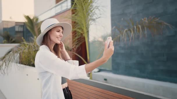 Giovane donna felice in abiti eleganti e cappello prendendo selfie su smartphone moderno mentre in piedi sulla strada. Viaggiatore femminile salvare i ricordi dalle vacanze estive. Donna felice scattare selfie su smartphone — Video Stock
