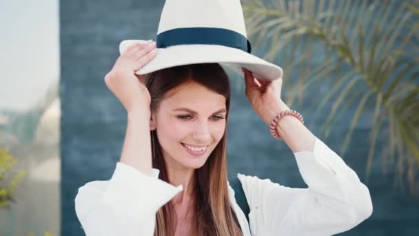 Hermosa mujer caucásica en gafas de sol y ropa de verano de pie en la calle y sonriendo en la cámara. Palmeras en el fondo. Clima soleado al aire libre. Mujer se pone un sombrero con estilo — Vídeo de stock