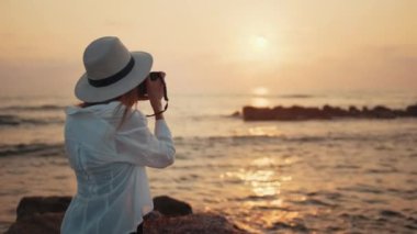 Şapkalı mutlu kadın denizde gün batımının fotoğrafını çekiyor.