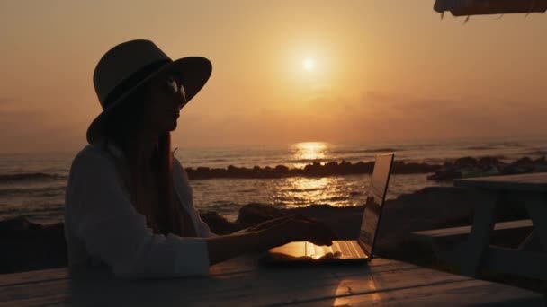 Жінка в літньому капелюсі сидить біля моря і використовує ноутбук — стокове відео