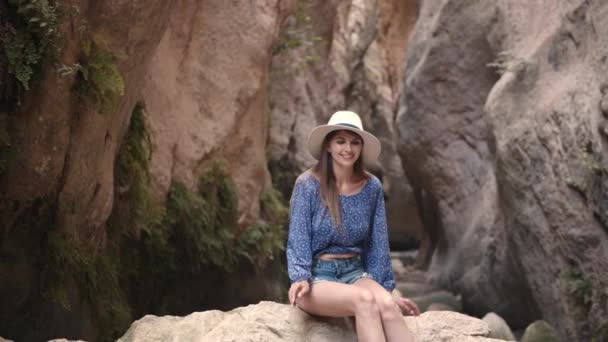 Oszałamiająca młoda kobieta siedząca na dużej skale pośród skał i uśmiechająca się do kamery. Biała kobieta w letnim kapeluszu i ubraniu. Ciesz się wakacjami. — Wideo stockowe