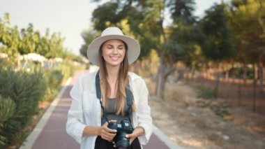 Genç fotoğrafçı gezgin kadın. Mutlu genç kadın yürürken fotoğraf çekmek için modern dijital kamera kullanıyor. Yazlık şapkalı ve elbiseli kadın turist, seyahatten kalan anıları saklıyor..
