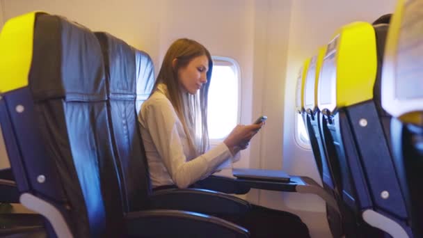 Mujer atractiva en ropa formal mirando a la cámara mientras está sentada cerca de la ventana del avión con un teléfono inteligente moderno en las manos. Viaje de negocios de una dama ocupada. Concepto de tecnología y asignación. — Vídeos de Stock