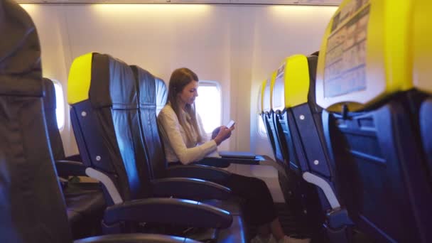 Vonzó nő formális ruhákban a kamerába néz, miközben a repülőgép ablaka mellett ül modern okostelefonnal a kezében. Egy elfoglalt hölgy üzleti útja. Technológia és hozzárendelés — Stock videók