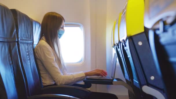 Tıbbi maske takan beyaz kadın ve uçuş sırasında kablosuz bilgisayarla çalışan resmi kıyafetler. Genç bayan uçağın penceresinin yanında oturuyor ve modern aletler kullanıyor. Uçakta dizüstü bilgisayar — Stok video