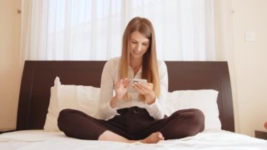 Otelde dinlenirken modern akıllı telefondan internette uzun saç sörfü yapan çekici genç bir kadın. Kafkasyalı mutlu kadın yatakta oturuyor ve cep telefonu kullanıyor. Dinlenirken akıllı telefon kullanan kadın