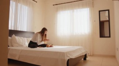 Yatakta elinde taze meyve tabağıyla oturan ve cep telefonuyla fotoğraf çeken mutlu genç bir kadın. Rahat bir otel odasında boş zamanın tadını çıkaran bir kadın gezgin. İnsanlar ve teknoloji kavramı.