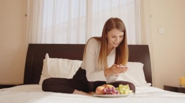 Genç ve mutlu bir kadın taze meyve tabağıyla yatakta oturuyor ve cep telefonuyla konuşuyor. Bayan gezgin rahat otel odasında boş zamanın tadını çıkarıyor. İnsanlar ve teknoloji kavramı.