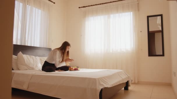 Szczęśliwa młoda kobieta siedząca na łóżku z talerzem świeżych owoców i robiąca zdjęcia telefonem komórkowym. Kobieta podróżnik korzystających z wolnego czasu w przytulnym pokoju hotelowym. Koncepcja ludzi i technologii. — Wideo stockowe