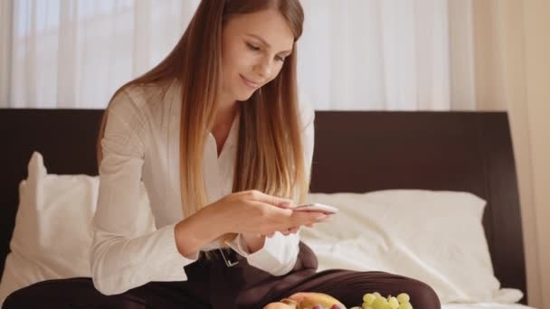 Genç ve mutlu bir kadın taze meyve tabağıyla yatakta oturuyor ve cep telefonuyla konuşuyor. Bayan gezgin rahat otel odasında boş zamanın tadını çıkarıyor. İnsanlar ve teknoloji kavramı. Yatakta oturan kadın — Stok video