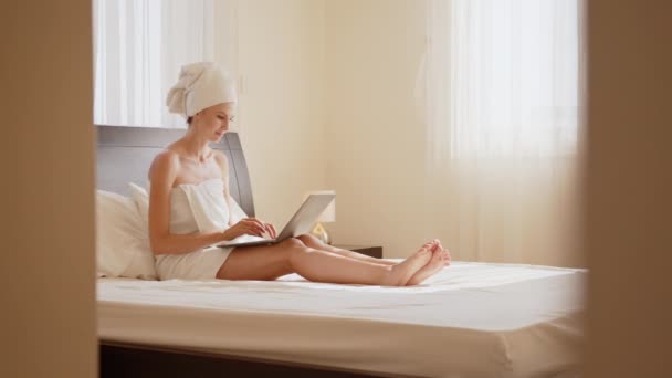Szczęśliwa biała kobieta korzystająca z bezprzewodowego laptopa podczas odpoczynku na łóżku w ręczniku kąpielowym. Kobieta podróżujący korzystających z czasu w wygodnym pokoju hotelowym z nowoczesnym urządzeniem. Szczęśliwa kobieta korzystająca z laptopa podczas odpoczynku na łóżku — Wideo stockowe