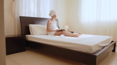 Banyo havlusu giymiş bir kadın otel odasında dizüstü bilgisayar kullanıyor. Beyaz kadın banyo havlusuyla yatarken kablosuz laptop kullanıyor. Dişi gezgin modern cihazlarla rahat otel odasında vakit geçiriyor..
