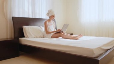 Beyaz kadın banyo havlusuyla yatarken kablosuz laptop kullanıyor. Dişi gezgin modern cihazlarla rahat otel odasında vakit geçiriyor. Banyo havlusu giymiş bir kadın otel odasında dizüstü bilgisayarla dinleniyor.