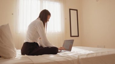 Rahat giyimli beyaz bir kadın bacak bacak üstüne atmış bir şekilde yatakta oturuyor ve kablosuz bilgisayarda yazıyor. Rahat otel odasında rahatlama sırasında modern teknolojiyi kullanmak. Yatakta oturan gülümseyen kadın.