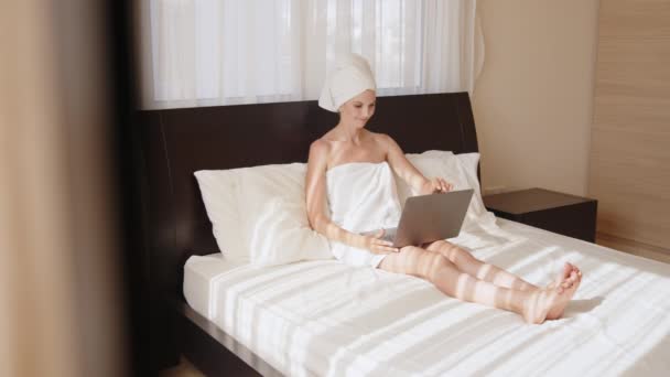 Młoda kobieta owinięta w ręcznik, leżąca na wygodnym łóżku i rozmawiająca podczas rozmowy wideo. Kobieta podróżująca za pomocą bezprzewodowego laptopa do rozmowy online z pokoju hotelowego. Kobieta ma rozmowę wideo na laptopie — Wideo stockowe