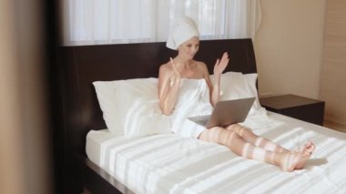 Havlu giymiş bir kadın otel odasında dizüstü bilgisayarla konuşuyor.