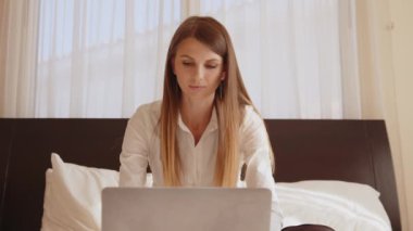Rahat bir yatakta oturan ve kablosuz bilgisayar kullanan rahat giyimli güzel bir genç kadın. Kafkasyalı kadın otel odasında modern aletlerle dinleniyor. Kadın rahat bir yatakta oturuyor ve kablosuz bilgisayar kullanıyor.
