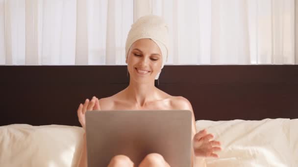 バスタオルを着た若い女性が居心地の良いベッドで休憩し、ビデオ通話のためにワイヤレスノートパソコンを使用しています。ホテルの部屋でオンライン通信をしている女性旅行者。ビデオ通話のためのラップトップを使用してバスタオルの女性. — ストック動画