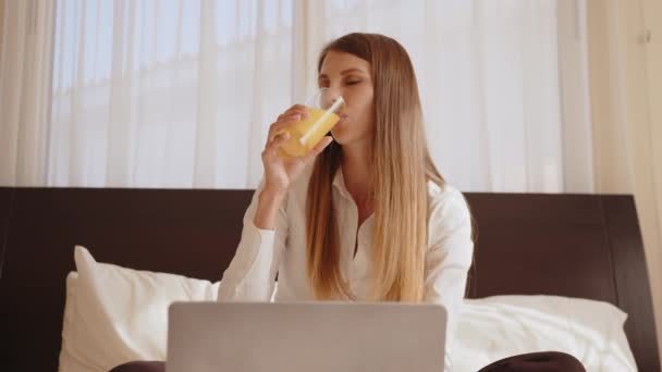 Biała młoda kobieta pisząca na laptopie, siedząc na łóżku ze świeżymi owocami i sokiem. Szczęśliwa pani ciesząca się wakacjami w przytulnym pokoju hotelowym. Kobieta pisze na laptopie siedząc na łóżku z owocami — Wideo stockowe