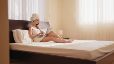 Banyo havlusuna sarılı güzel bir kadın dizüstü bilgisayarla yatağa uzanmış ve akıllı telefonu elinde tutuyor. Modern teknolojili yaşam tarzları. Bir kadın banyodan ya da duştan sonra otelde tatilde çalışır.
