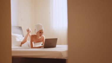 Otel odasında dizüstü bilgisayar kullanarak banyo havlusu içinde gülümseyen kadın.