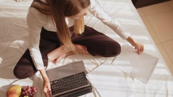 Kobieta siedzi na łóżku z dokumentami i pisze na laptopie — Wideo stockowe