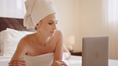 Banyo havlusundaki kadın rahat yatakta taşınabilir laptop kullanıyor.