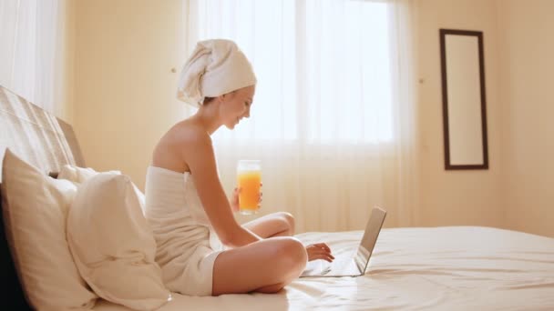 Kobieta owinięta w ręcznik przy użyciu laptopa i picia soku na łóżku — Wideo stockowe