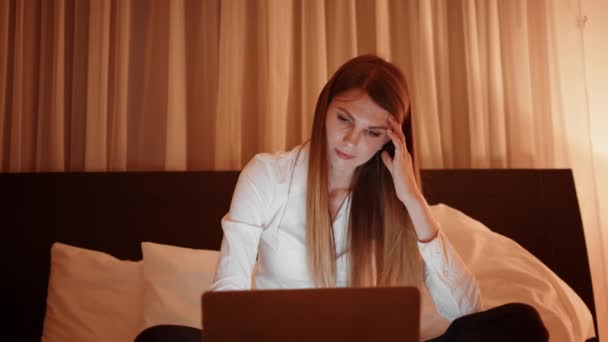 Yatakta oturup dizüstü bilgisayarla uğraşan yorgun bir kadın. — Stok video
