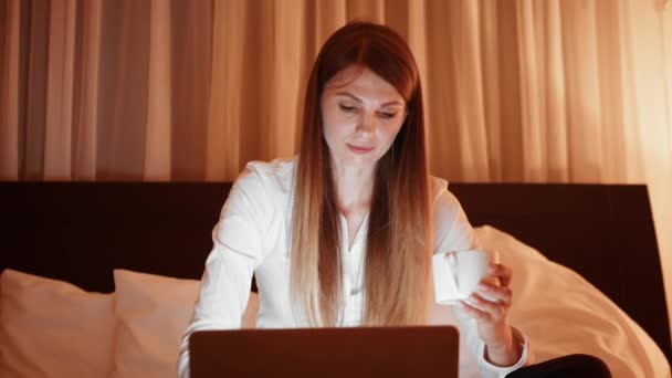 Mujer joven bebiendo café y utilizando el ordenador portátil en la habitación del hotel — Vídeos de Stock