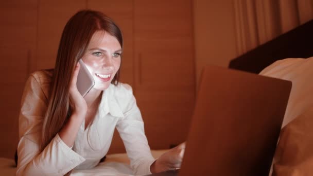 Mulher falando no celular e digitando no laptop na cama — Vídeo de Stock