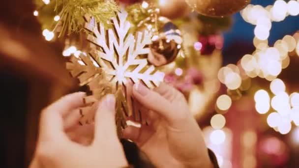 Großaufnahme von Händen, die den Weihnachtsbaum mit Kugeln vor dem Hintergrund heller festlicher Lichter schmücken. Kinder schmücken einen Weihnachtsbaum. Weihnachtsbaum schmücken, Spielzeug auf Ast. — Stockvideo