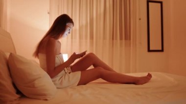 Banyo havlusu giymiş genç bir kadın yatağa uzanıyor ve cep telefonu kullanıyor.