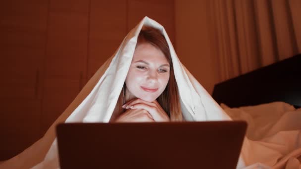 Kvinna som sitter på sängen under vit filt och använder laptop — Stockvideo